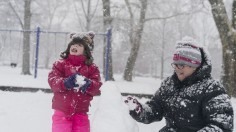 Girl catching snowflakes with her tongue while she makes a snowman with her mother 