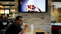 eating, Customer Steven Price sits at a table near a HDTV screen showing the new McDonald's Channel featuring a commercial about McCafe drinks at a McDonald's restaurant.