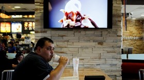 eating, Customer Steven Price sits at a table near a HDTV screen showing the new McDonald's Channel featuring a commercial about McCafe drinks at a McDonald's restaurant.